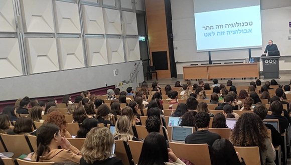 הרצאה באולם ומצגת שבה כתוב: "טכנולוגיה זזה מהר, אבולוציה זזה לאט".